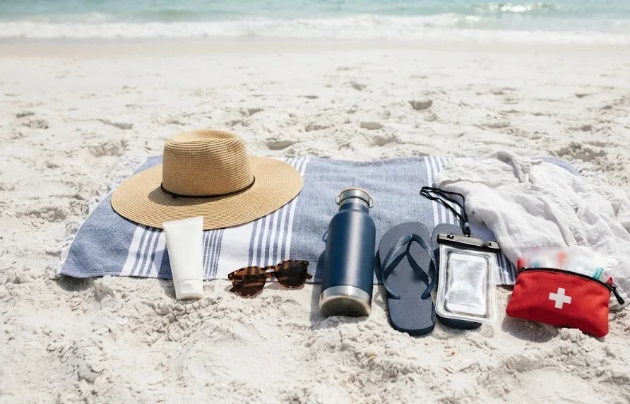 Sun hat, sunscreen, sunglasses, water bottle, flip flops, phone pouch and first aid kit on a towel