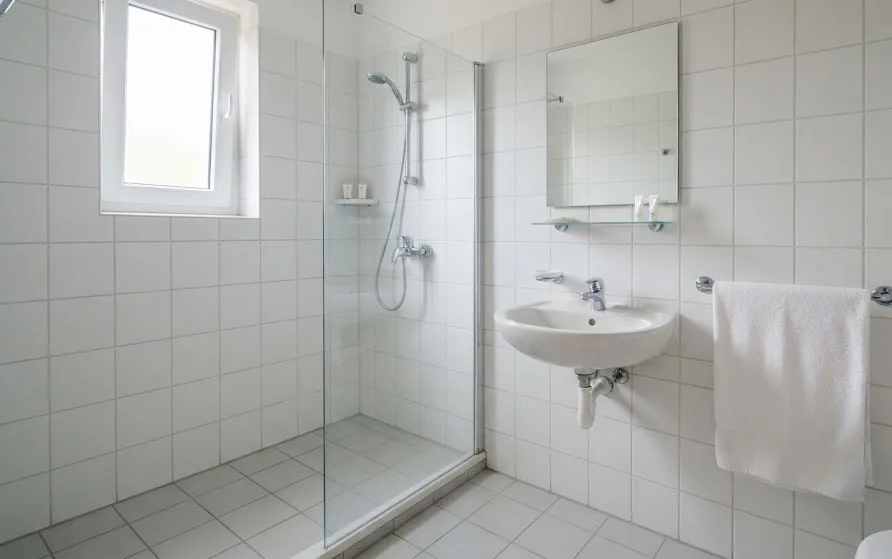 Small white-tiled hotel bathroom with a walk-in shower, basin, mirror and folded towel