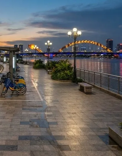 Riverside promenade with a bike-share dock and a lit dragon bridge at dusk