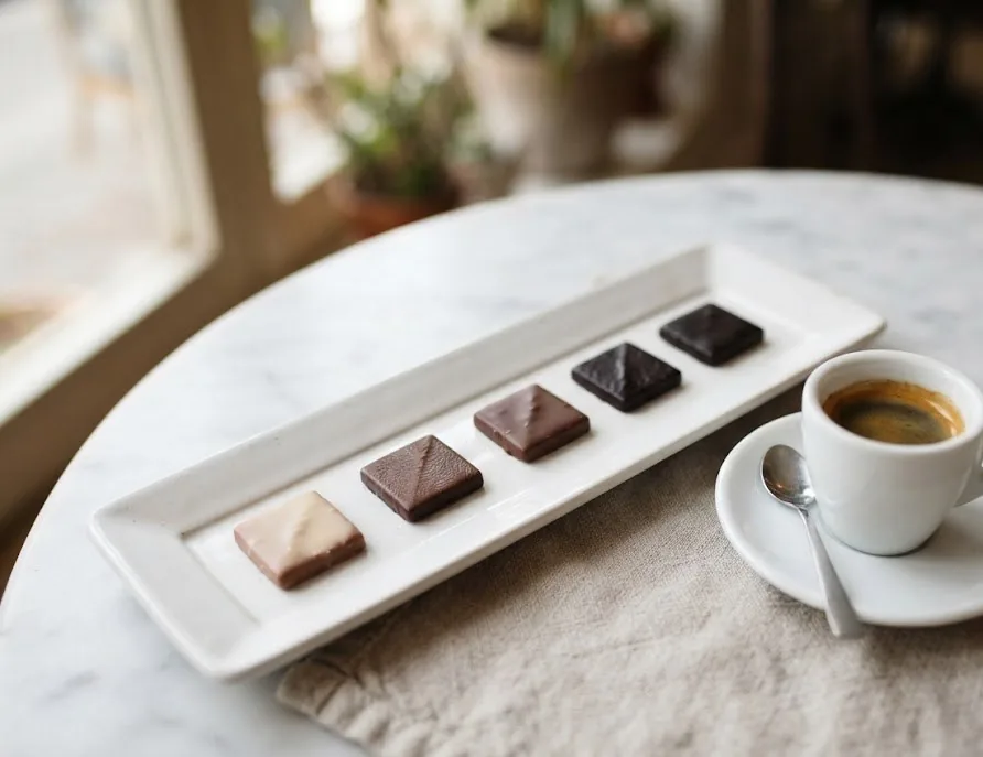 Five squares of chocolate from pale to dark on a white plate beside a cup of black coffee