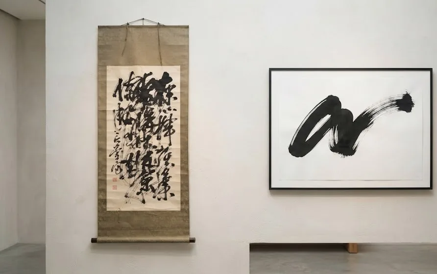 Aged hanging scroll of dense brushwork beside a framed modern piece with a single sweeping stroke