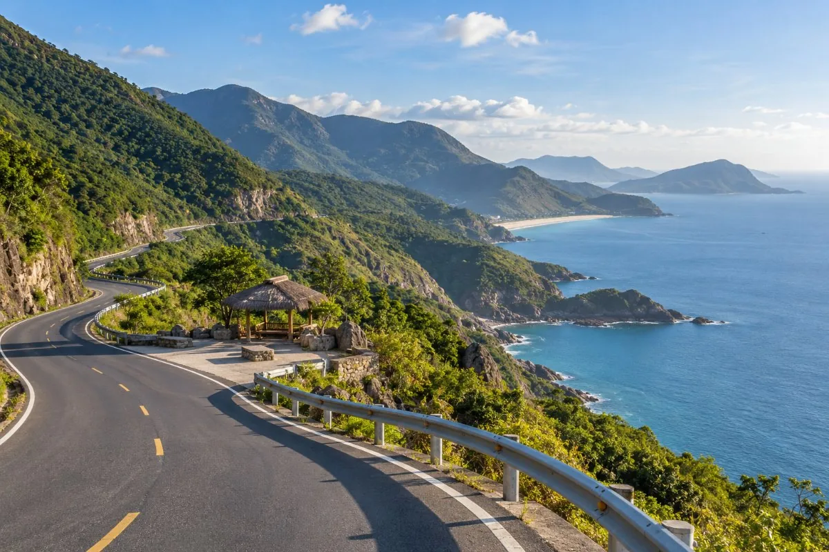 Winding mountain pass road with guard rails and a thatched rest stop above the sea