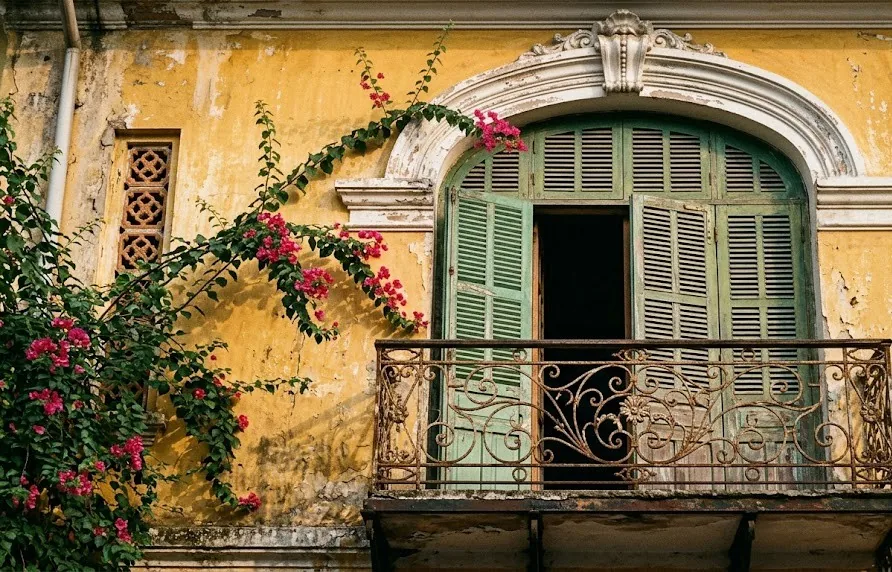 Arched window with green louvred shutters, plaster mouldings and wrought iron balcony on an old yellow wall