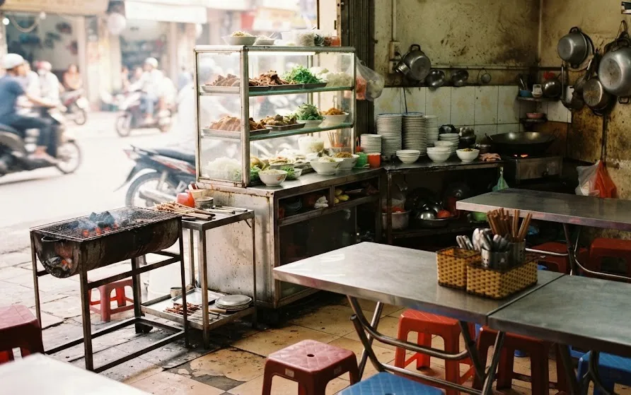Small street-side eatery with stainless steel tables, plastic stools, a food display cabinet and a charcoal grill