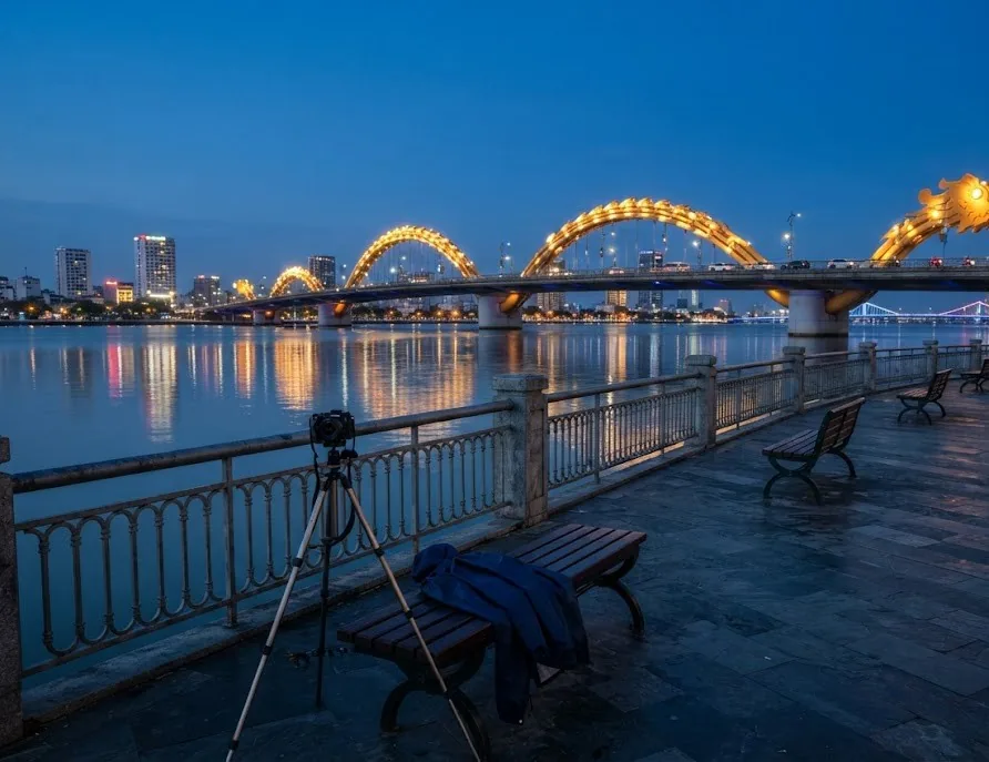 Riverside promenade with a tripod and bench facing the lit dragon bridge at blue hour
