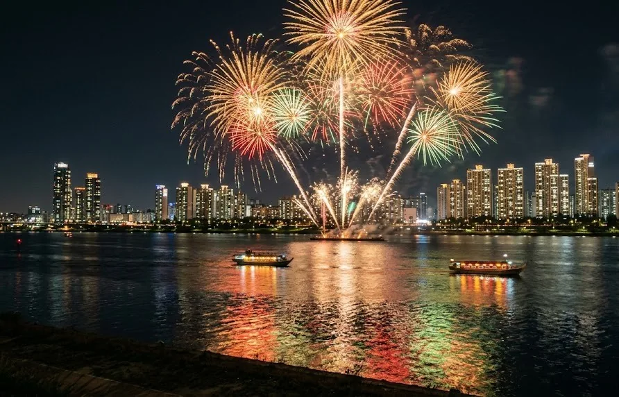 Gold and green fireworks bursting over a wide city river with small lit boats at night
