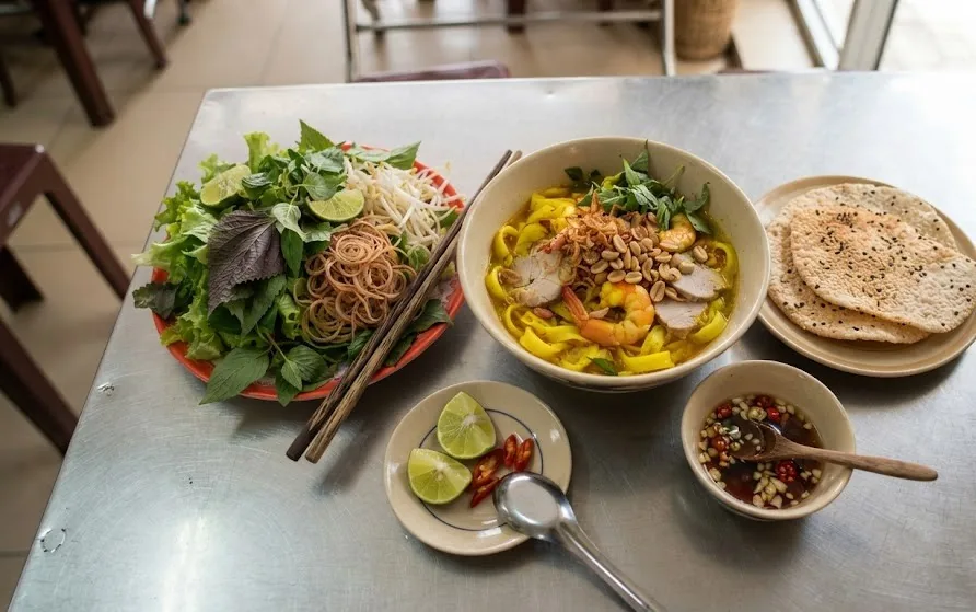 Turmeric noodle bowl served with a platter of fresh greens, sesame rice crackers, lime and chilli