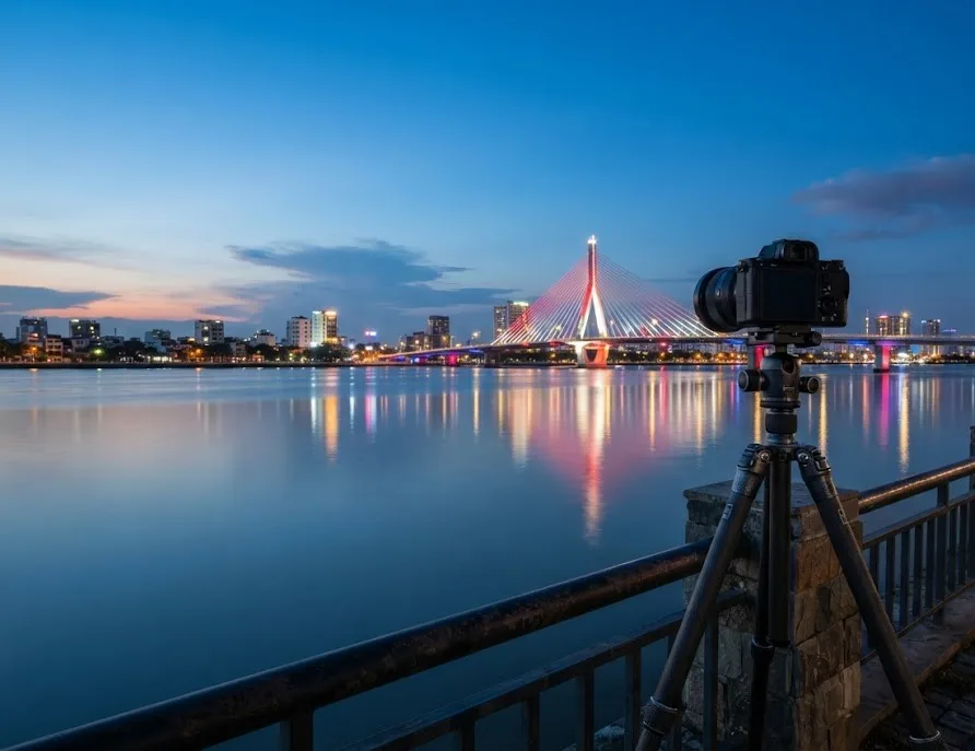 Camera on a tripod capturing a lit cable-stayed bridge over a calm river at blue hour