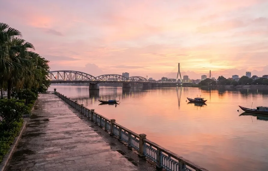 Riverside promenade at sunrise with an arched bridge, a cable stayed bridge and fishing boats