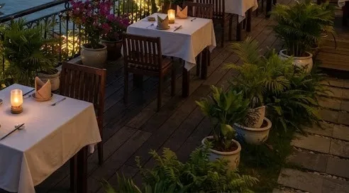 Candlelit tables with white cloths and potted plants on a riverside restaurant terrace