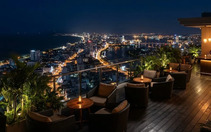Rooftop bar with rattan seating and candles overlooking a lit coastal city and dark sea at night