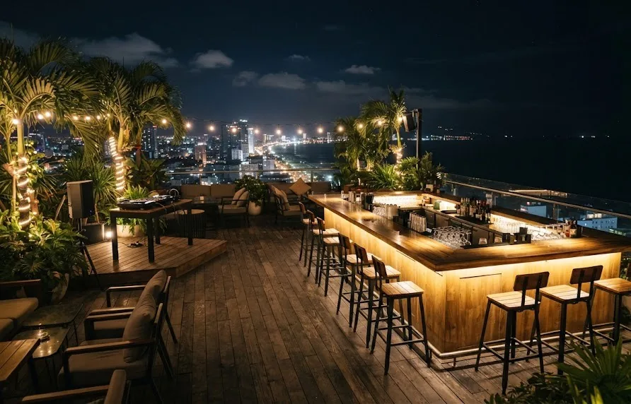Rooftop bar with a lit counter, stools and palms overlooking city lights and the sea