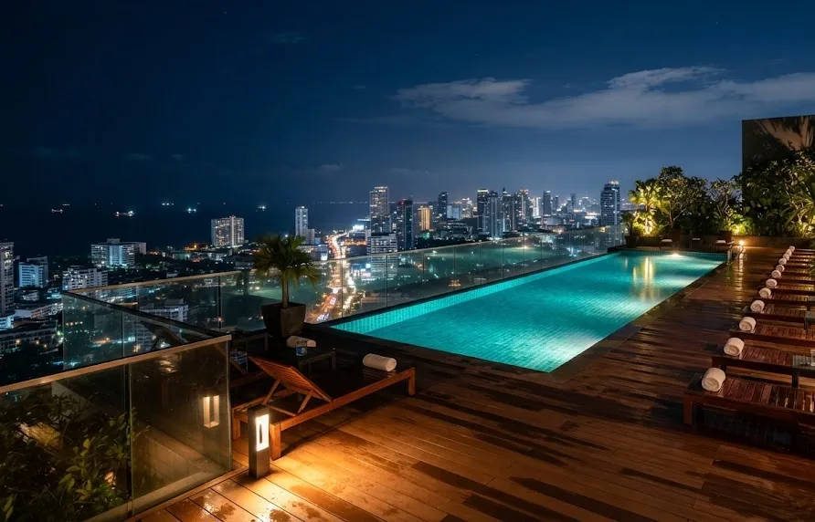 Turquoise rooftop pool glowing at night above a lit coastal city skyline and dark sea