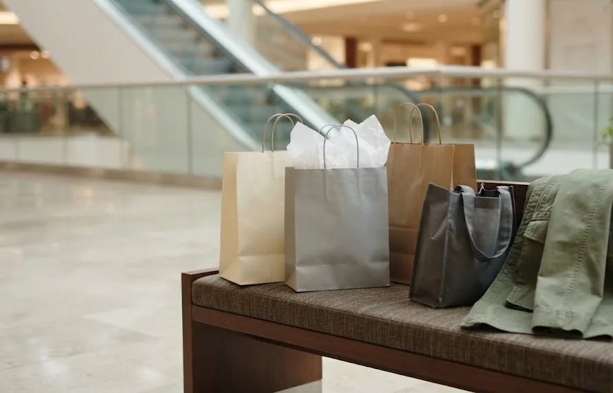 Plain paper shopping bags and a tote on a mall bench with an escalator blurred behind