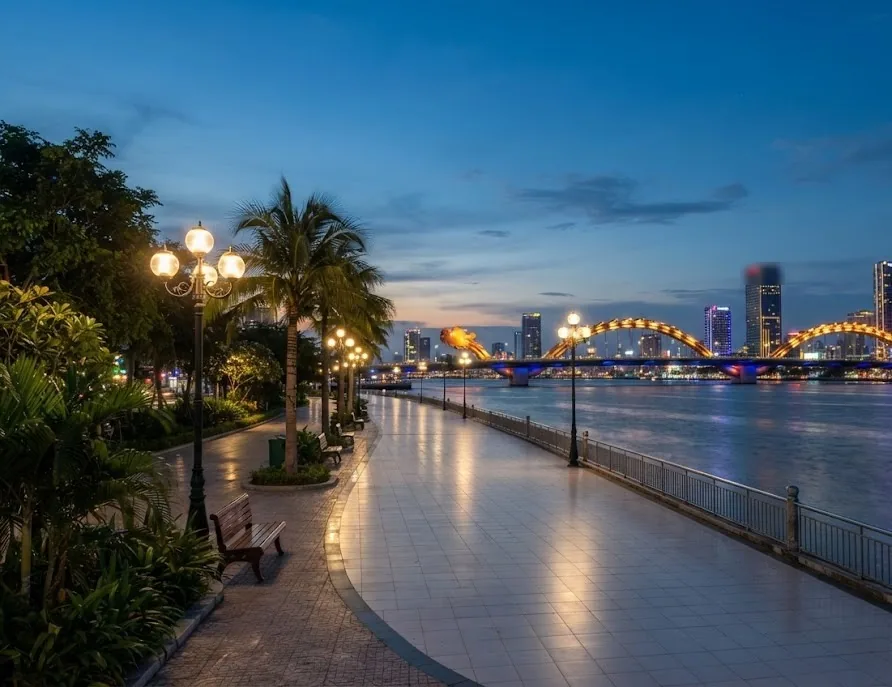 Well-lit riverside promenade with lamps, benches and a lit dragon bridge at dusk